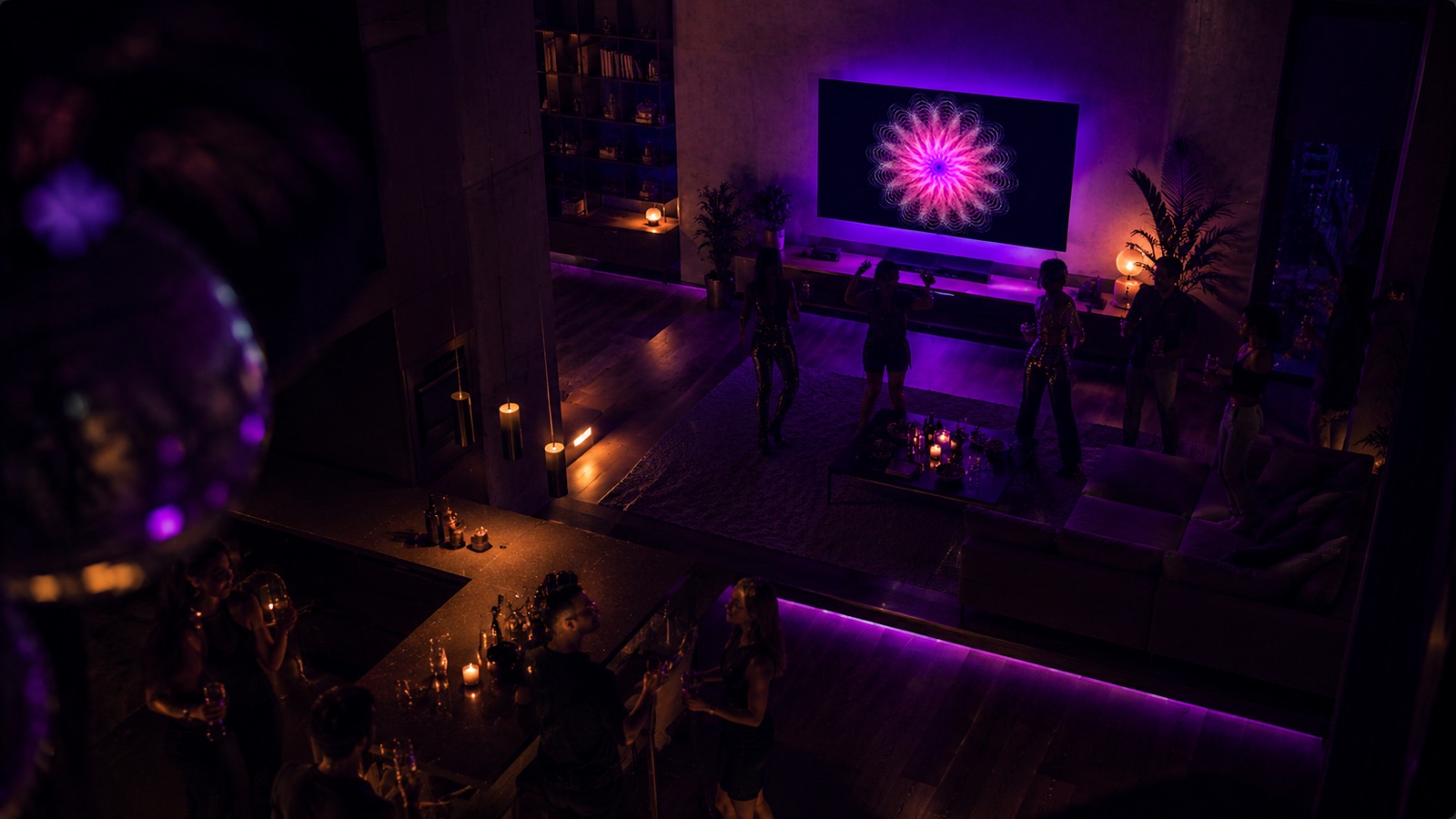 Overhead view of a multi-level loft party at night — a wall TV runs a glowing magenta-pink Spiralyst Lab fractal while guests dance in front of it, a disco ball catches the colored light in the foreground, and more guests mingle at a candle-lit bar below.